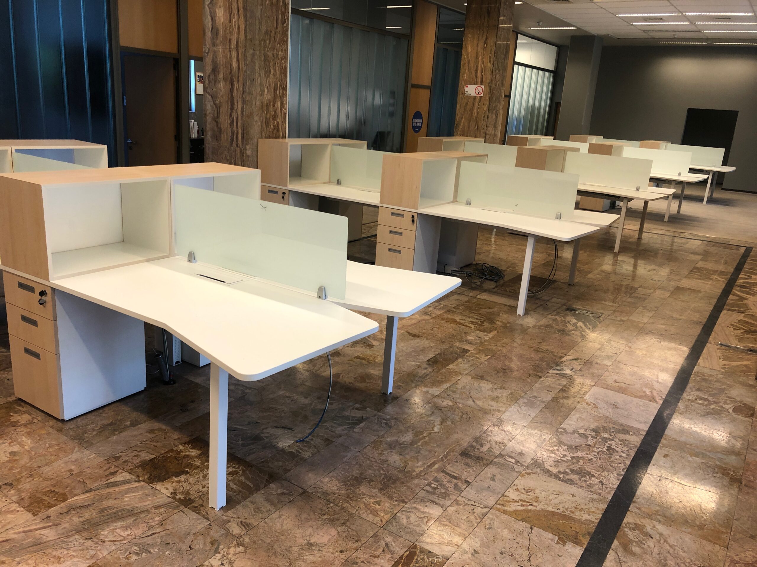 Conjunto de mesas blancas con separadores y cajoneras sobre un piso de tonos cálidos.