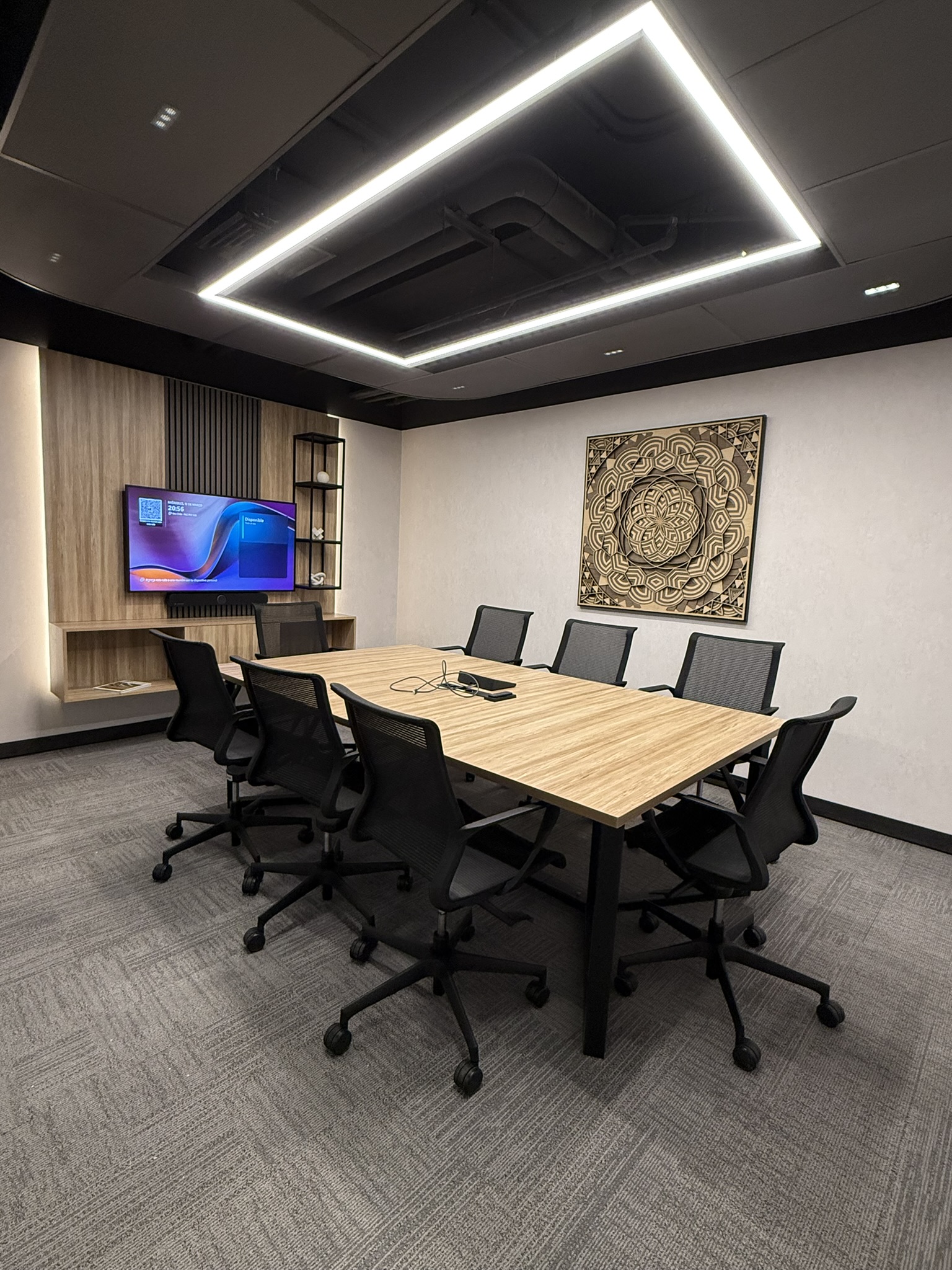 Sala de reunión con mesa de madera clara, sillas negras y una obra decorativa en la pared.