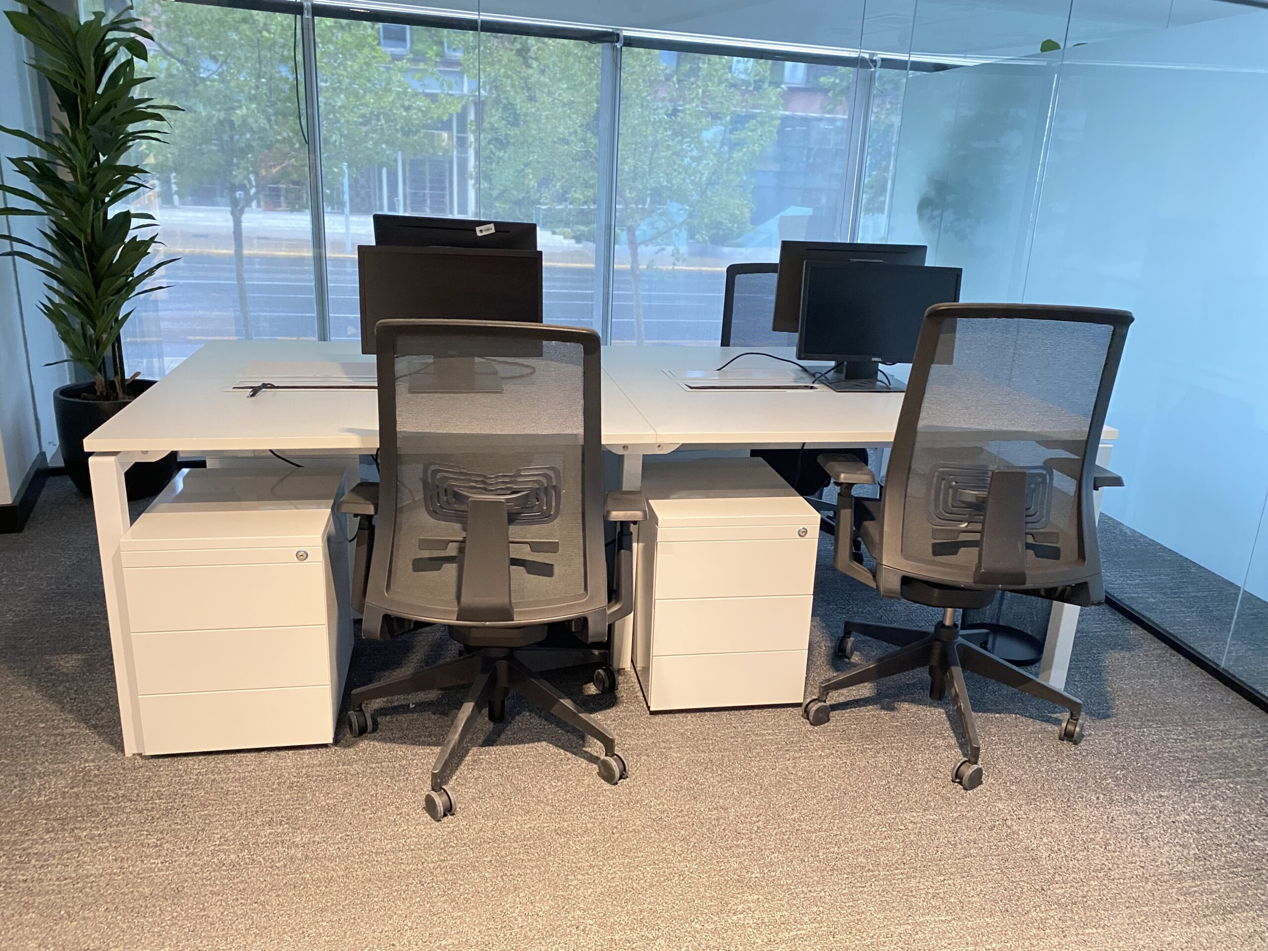 Puestos de trabajo blancos con cajoneras y sillas de respaldo de malla junto a una ventana.